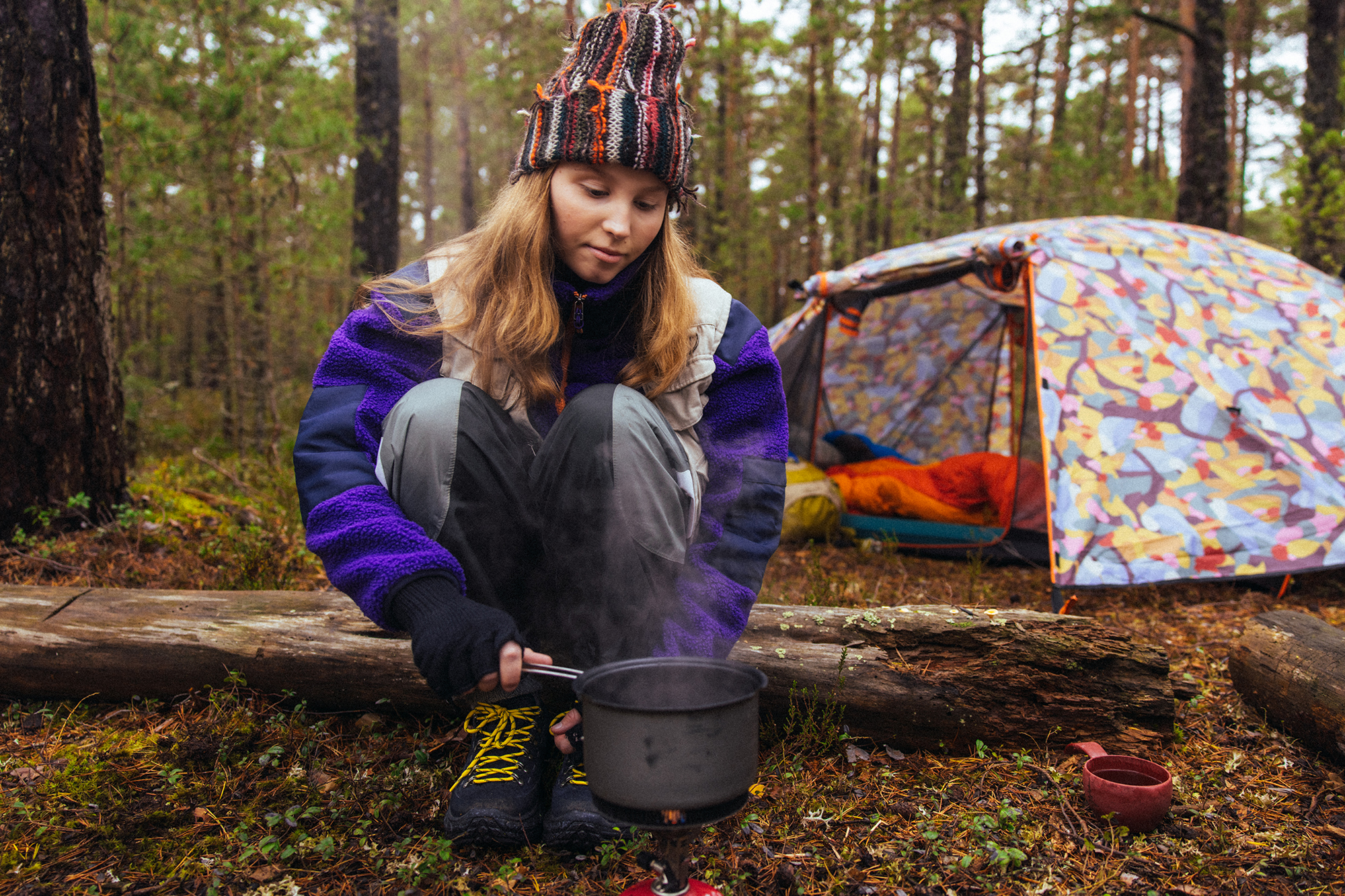 Tyttö keittää vettä kattilassa ja teltta on taustalla metsämaisemassa.