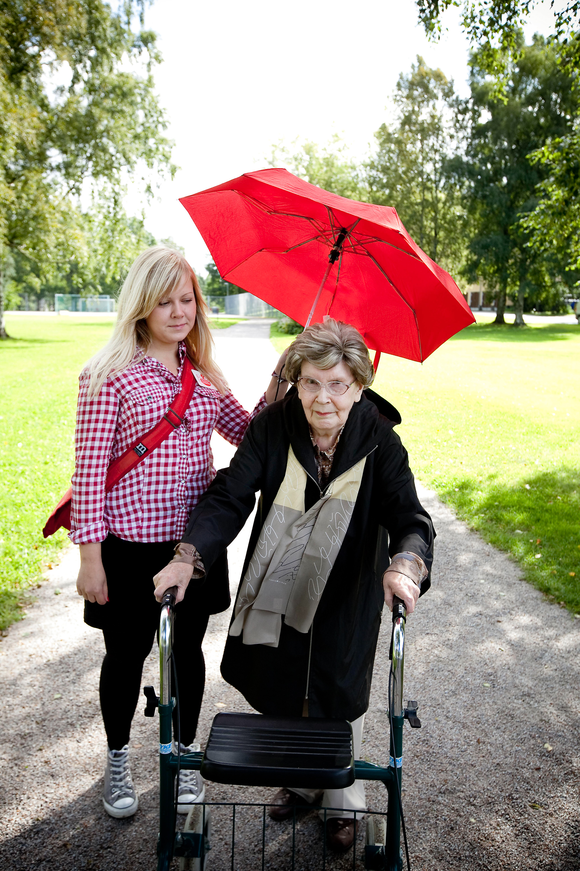 Nainen pitää punaista sateenvarjoa rollaattorilla kulkevan iäkkäämmän naisen päällä.