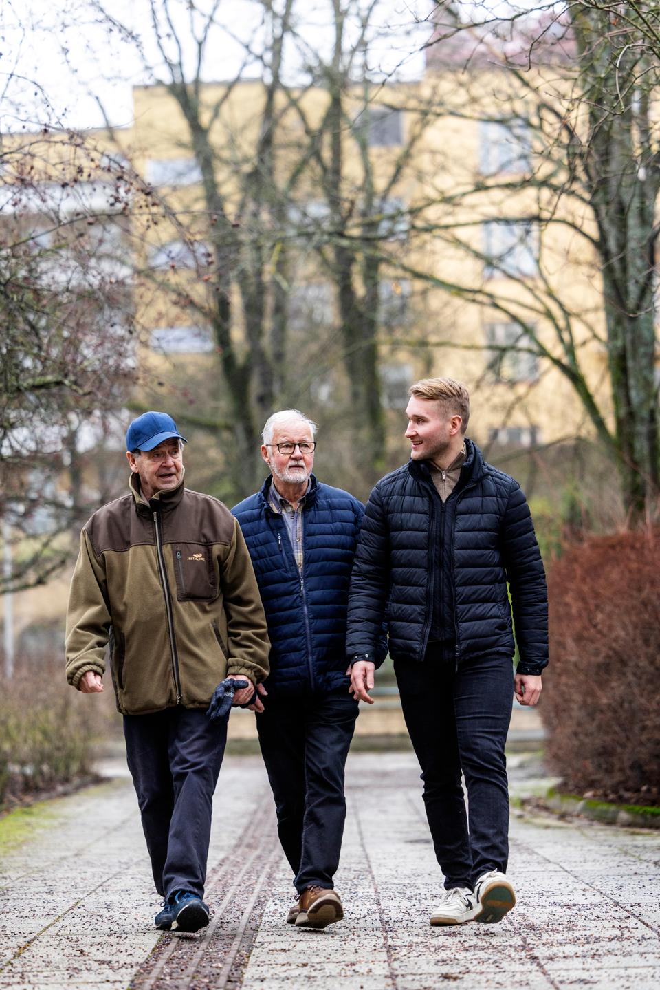 Kolme ihmistä, joukossa kaksi vanhempaa ja yksi nuorempi, kulkee rinnakkain piha-alueella.